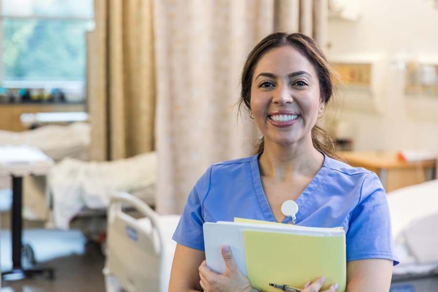 Cheerful nurse in scrubs holding paperwork in folder standing in patient room
