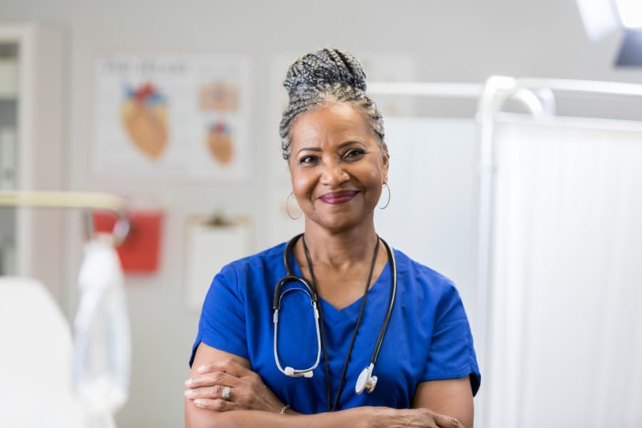 Nurse smiling in exam room with arms folded wearing scrubs and stethoscope 