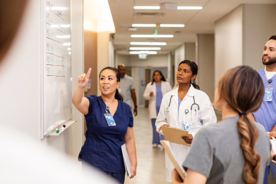 Nurse in scrubs gesturing to whiteboard in hospital with care team